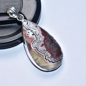 以下が含まれることがあります： 銀色のベイルが付いた、涙の形をしたペンダント。ペンダントは、赤、白、茶色などの色が混ざった磨かれた石が特徴です。石は銀色のベゼルにセットされています。