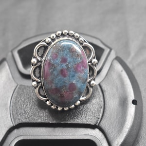 May include: Oval gemstone ring with a silver setting. The stone features a blue-green base with red inclusions. The ring has a decorative border with small beaded accents.