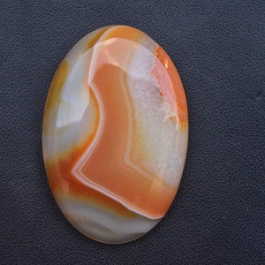 May include: Oval-shaped polished gemstone with bands of orange, white, and yellow. The stone has a smooth, glossy surface and a natural, organic appearance. The gemstone is a cabochon, suitable for jewelry making.