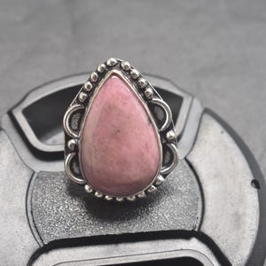 Op de afbeelding: Een zilveren ring met een druppelvormige roze edelsteen. De ring heeft een sierlijke zilveren zetting met decoratieve details rond de steen. De edelsteen heeft een zachtroze kleur, tegen een grijze achtergrond.