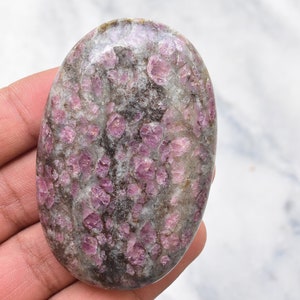 May include: An oval-shaped polished gemstone featuring a mottled pattern of vibrant magenta and deep pink crystalline inclusions embedded within a translucent grey and white matrix, showcasing a smooth, glossy surface texture throughout.