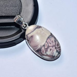 以下が含まれることがあります： シルバーのベイルが付いた楕円形のペンダント。クリーム色と紫色の模様の研磨された石が特徴です。ペンダントは白と黒の背景に配置されています。