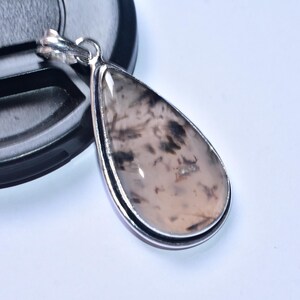 以下が含まれることがあります： 銀色のフレームが付いた、涙の形をしたペンダント。ペンダントの石は、明るいベージュ色のベースに濃い茶色と黒のインクルージョンがあります。ペンダントは、黒いオブジェクトの隣の白い表面にあります。