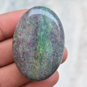 Può includere: Una gemma ovale con una superficie lucida. La pietra presenta un mix di colori, tra cui verde, viola e grigio. La gemma è tenuta in una mano, mostrando le sue dimensioni e la sua consistenza. La superficie della pietra ha un effetto scintillante.