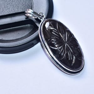 以下が含まれることがあります： 表面に花の模様が彫られた、楕円形の黒いペンダント。ペンダントは銀色の金属で縁取られ、お揃いのベイルが付いています。白い表面にペンダントが表示されています。