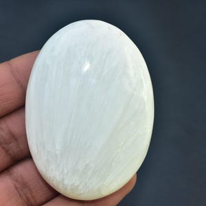 May include: A smooth, oval-shaped, pale green gemstone. The stone is held in a hand, and the surface has subtle, natural veining. The gemstone is approximately 3 inches long.