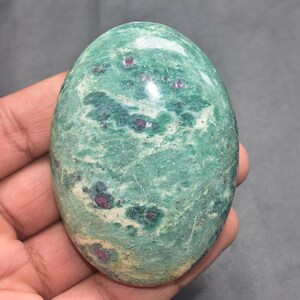 Op de afbeelding: Een ovaalvormige gepolijste edelsteen met een gevlekt groen oppervlak met duidelijke witte vlekken en kleine, ingebedde diepmagenta-kleurige insluitingen door de hele steen. Het oppervlak heeft een gladde, glanzende afwerking die licht reflecteert.