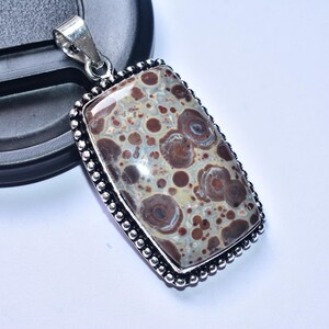 以下が含まれることがあります： 銀色のバチカンと模様入りの石が付いた長方形のペンダント。石は、茶色、ベージュ、青灰色の色が混ざり合い、斑点模様のデザインになっています。ペンダントは、ビーズの銀色の縁で縁取られています。