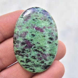 Könnte beinhalten: Ovaler Zoisit-Edelstein mit polierter Oberfläche. Der Stein weist ein gesprenkeltes Muster aus grünen, schwarzen und tiefroten Farbtönen auf. Der Edelstein wird in einer Hand gehalten und zeigt seine natürlichen Farben und Texturen.