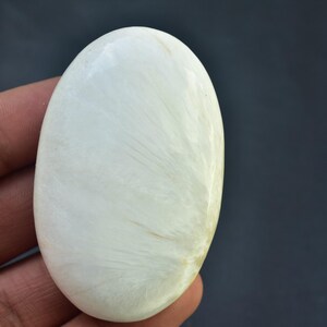 May include: A smooth, oval-shaped, off-white gemstone with subtle, feathery patterns. The polished stone is held in a hand, showcasing its natural texture and color against a dark background. The stone's shape is reminiscent of a cabochon.