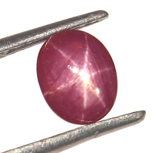 Pode incluir: Uma gema de rubi estrela rosa com uma estrela de seis pontas no centro, segurada por pinças.