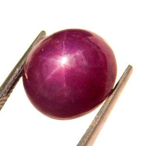 May include: A deep red star ruby gemstone with a star-shaped reflection in the center. The ruby is round and smooth, and it is held in place by a pair of tweezers.