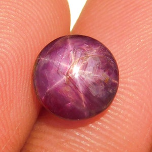 May include: A round, polished gemstone with a deep magenta hue. The gem features a six-rayed star pattern across its surface, with lighter streaks radiating from the center. The stone is held between fingers against a white background.