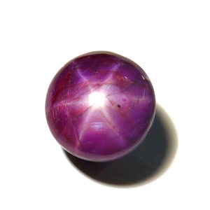 May include: A round, polished, purple gemstone with a six-rayed star effect. The gem has a smooth, reflective surface and a bright, central point of light. The star pattern is clearly visible across the surface of the stone.