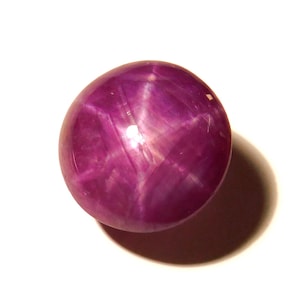 Può includere: Una gemma rotonda e lucidata di un profondo colore magenta. La gemma mostra un effetto a stella sulla sua superficie, con la luce che si riflette in un motivo a sei raggi. La pietra è posta su uno sfondo bianco, proiettando un'ombra.