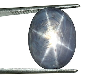 Könnte beinhalten: Ein blau-grauer ovaler Edelstein, gehalten von einer Metallpinzette, mit einem sechsstrahligen Sternmuster vor weißem Hintergrund. Der Sterneffekt entsteht durch Lichtreflexionen an Einschlüssen im Inneren. Der Edelstein ist poliert und glatt.