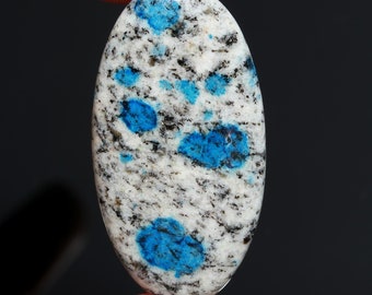 May include: Oval-shaped K2 stone cabochon. The stone features a white base with speckled black and gray patterns, and vibrant blue orb-like inclusions. The polished surface reflects light, highlighting the stone's natural beauty. The cabochon is held by a finger.