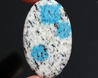 May include: Oval-shaped k2 stone with a polished surface. The stone features a white base with black speckles and vibrant blue patches. The gemstone is held between two fingers against a dark background.