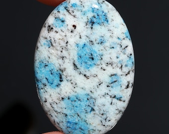 May include: An oval-shaped gemstone with a polished surface. The stone features a white base with blue and black speckled patterns. The blue patterns are clustered, creating a unique visual texture. The gemstone is held between two fingers.