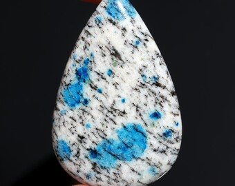 May include: A teardrop-shaped K2 stone, featuring a white base with black and blue speckles and patches. The polished gemstone is held by a finger against a dark background. The stone's unique pattern makes it suitable for jewelry.