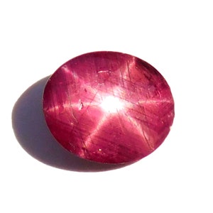 Pode incluir: Uma pedra preciosa de rubi oval polida com efeito de estrela. A gema é de cor vermelho-rosada profunda, com um padrão de estrela de seis raios irradiando do centro. A pedra está sobre um fundo branco.