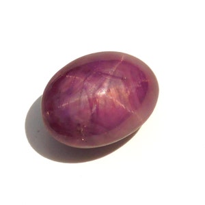Può includere: Una gemma lucida, di forma ovale, di un profondo colore rosso-violaceo. La gemma ha una superficie liscia e riflettente e un sottile motivo a forma di stella. La pietra è posta su uno sfondo bianco, con un'ombra morbida.