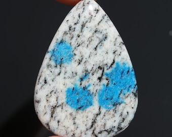 May include: A teardrop-shaped K2 stone, featuring a white base speckled with black and vibrant blue patches. The polished gemstone is held between fingers against a dark background, showcasing its natural patterns and colors. This unique stone is ideal for jewelry making.