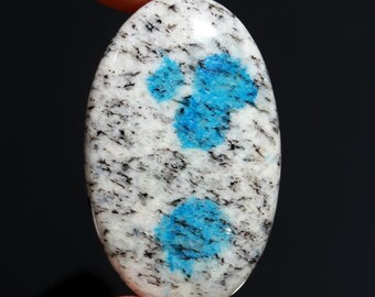 May include: An oval-shaped K2 stone with a polished surface. The stone features a white base speckled with black and two vibrant blue patches. The stone is held by a finger against a black background.