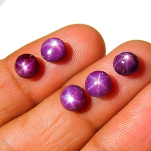 May include: Four purple star ruby cabochons with a star-like pattern in the centre. The stones are round and have a smooth, polished surface.