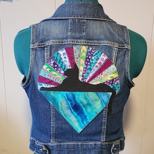 May include: Blue denim vest with a colorful, artistic design on the back. The design features a black silhouette of a cat, a colorful sunburst, and a blue and green water-like pattern. The vest is displayed on a teal dress form.