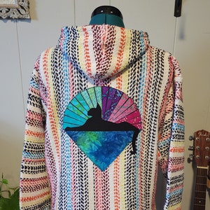 Puede incluir: Una sudadera con capucha tejida multicolor con un diseño de silueta de gato en la espalda. La sudadera presenta rayas verticales en varios colores, incluyendo rosa, azul, amarillo y negro. El diseño del gato está sobre un fondo en forma de abanico.