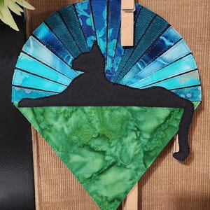 Peut inclure: Décoration murale artisanale sur le thème du chat. Le motif présente une silhouette de chat noir se prélassant sur un fond en tissu vert et bleu. Le fond est en forme d'éventail avec des nuances de bleu et de turquoise. Le tout est maintenu par une pince à linge en bois.