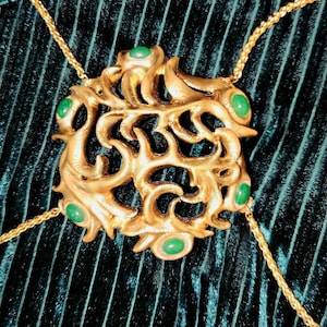 Könnte beinhalten: Eine goldfarbene Körperkette mit einem verzierten, durchbrochenen Anhänger. Der Anhänger ist mit grünen Edelsteinen besetzt und mit zarten Goldketten verbunden. Der Schmuck wird vor einem dunkelgrünen, strukturierten Hintergrund präsentiert.