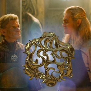 Può includere: Due uomini stanno in piedi in una stanza scarsamente illuminata. L'uomo a sinistra indossa una veste bordeaux con ricami dorati. L'uomo a destra indossa una camicia marrone chiaro e ha lunghi capelli biondi. Un grande oggetto metallico ornato di colore dorato si trova al centro dell'immagine.