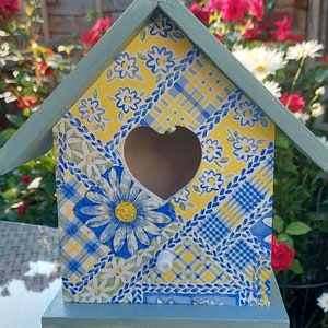 May include: A blue and yellow birdhouse with a heart-shaped opening. The birdhouse is decorated with a floral pattern and is sitting on a glass jar.