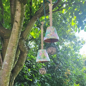 May include: Three hanging garden ornaments. Each has a floral-painted terracotta pot suspended by a rope, with a wire cage holding a round, textured object below. The decorations hang from a green metal hook attached to a tree branch, with green foliage in the background.