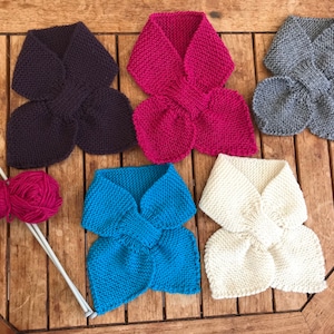 May include: A selection of hand-knitted scarves in shades of purple, fuchsia, grey, turquoise, and cream. The scarves are arranged on a wooden surface, with knitting needles and a ball of fuchsia yarn also visible.