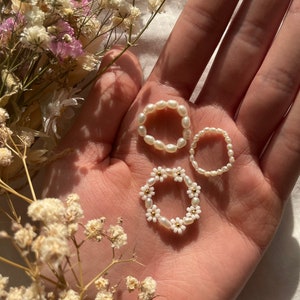 Può includere: Tre delicati anelli di perle e fiori. Gli anelli sono bianchi e presentano piccole perle e dettagli di fiori bianchi. Gli anelli sono mostrati su una mano.