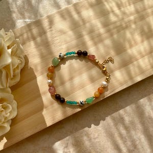 Puede incluir: Un brazalete de cuentas de colores con cuentas verdes, naranjas, marrones, blancas y rosas. El brazalete tiene un cierre dorado.