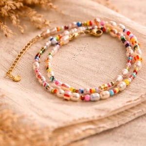 May include: A delicate necklace featuring freshwater pearls and colorful glass beads. The necklace has a gold-toned chain and clasp. The beads are in shades of red, yellow, blue, and green. The necklace is displayed on a beige cloth.