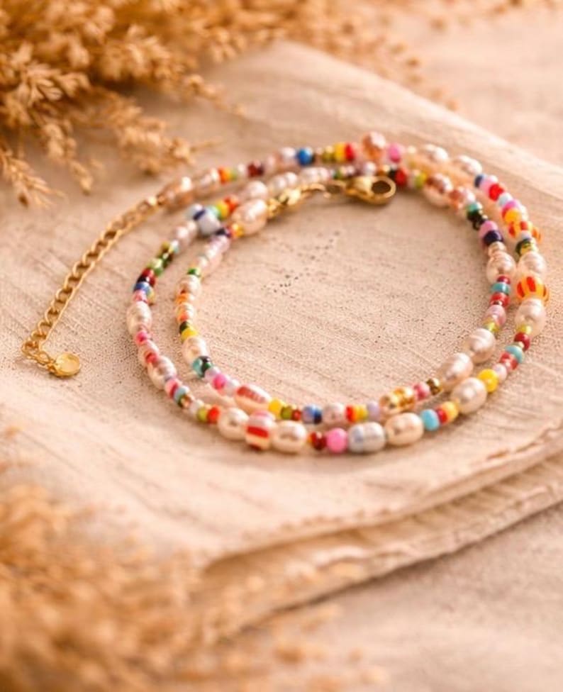 May include: A delicate necklace featuring freshwater pearls and colorful glass beads. The necklace has a gold-toned chain and clasp. The beads are in shades of red, yellow, blue, and green. The necklace is displayed on a beige cloth.