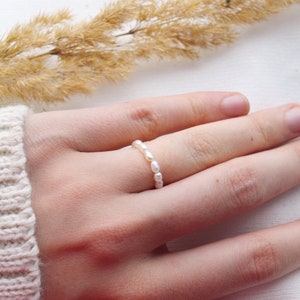 Puede incluir: Un delicado anillo con una serie de pequeñas perlas blancas. El anillo se lleva en un dedo, mostrando su elegante diseño. Las perlas están espaciadas uniformemente, creando un aspecto clásico y atemporal. Una joya.
