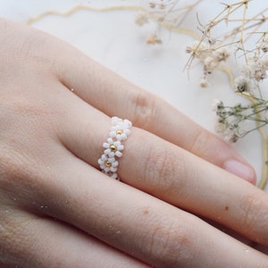 May include: A white daisy flower ring with gold accents. The ring is made of small beads and is worn on a finger.
