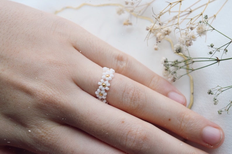 May include: A white daisy flower ring with gold accents. The ring is made of small beads and is worn on a finger.