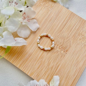 Könnte beinhalten: Ein zarter goldener Ring mit einer Reihe kleiner weißer Perlen. Der Ring wird auf einer Holzoberfläche mit weißen Blumen präsentiert.