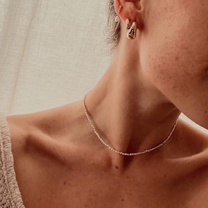 Könnte beinhalten: Eine zarte Perlenkette und goldene Ohrringe werden gezeigt. Die Halskette besteht aus kleinen, runden Perlen. Die Ohrringe sind goldfarben und haben eine einzigartige, organische Form. Der Schmuck wird getragen.