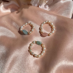 Könnte beinhalten: Drei Ringe mit Süßwasserperlen und Edelsteinakzenten. Ein Ring hat einen hellblauen Edelstein, ein anderer einen rosa Edelstein und der dritte einen grünen Edelstein. Die Ringe liegen auf einem rosa Satin.