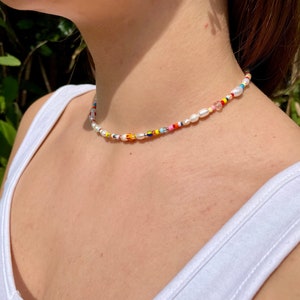 May include: A colorful beaded necklace with a mix of white pearls and small, vibrant beads in various colors. The necklace is worn around the neck, with a white tank top in the background.
