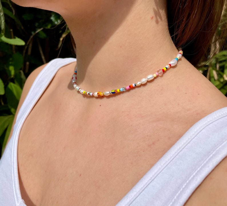 May include: A colorful beaded necklace with a mix of white pearls and small, vibrant beads in various colors. The necklace is worn around the neck, with a white tank top in the background.