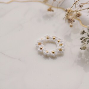 May include: A white beaded ring with gold accents, shaped like a daisy flower, on a white marble surface.
